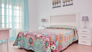 A bedroom with a colorful bedspread, two bedside tables with lamps, a desk, and a window with sheer curtains.