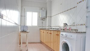 A kitchen with white tiled walls, wooden cabinets, a washing machine, and a window.