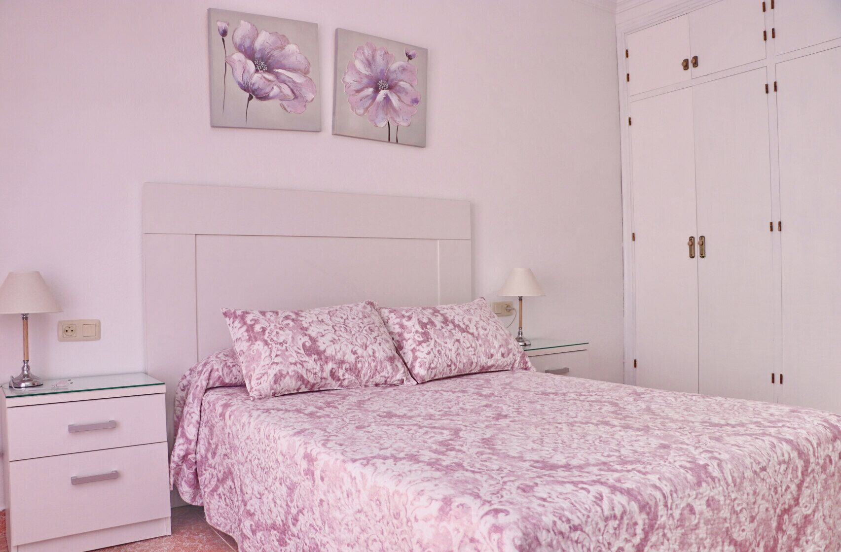 A bedroom with a bed, two nightstands, a dresser, a wardrobe, and two floral paintings on the wall.