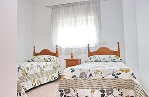 Two single beds with wooden headboards and floral-patterned bedding, a bedside table, and a large window with sheer curtains.