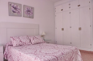 Ein Schlafzimmer mit einem Bett, zwei an der Wand befestigten floralen Bildern, einem Nachttisch, einer Lampe und einem großen Kleiderschrank.