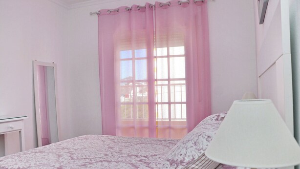 A bedroom with a bed, a bedside lamp, a window with pink curtains, and a mirror. - Apartment Violeta on the second floor (Conil de la Frontera)
