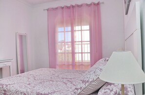 Een slaapkamer met een bed, een bedlamp, een raam met roze gordijnen en een spiegel.