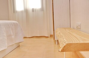 Quarto com uma cama, um banco de madeira e uma parede branca com um interruptor de luz.
