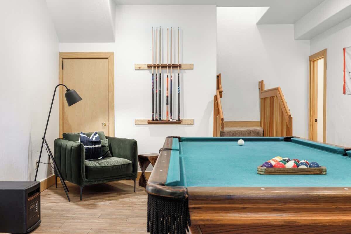 Une table de billard avec des billes, un fauteuil vert, une lampe et un porte-ques en bois contenant des queues de billard.