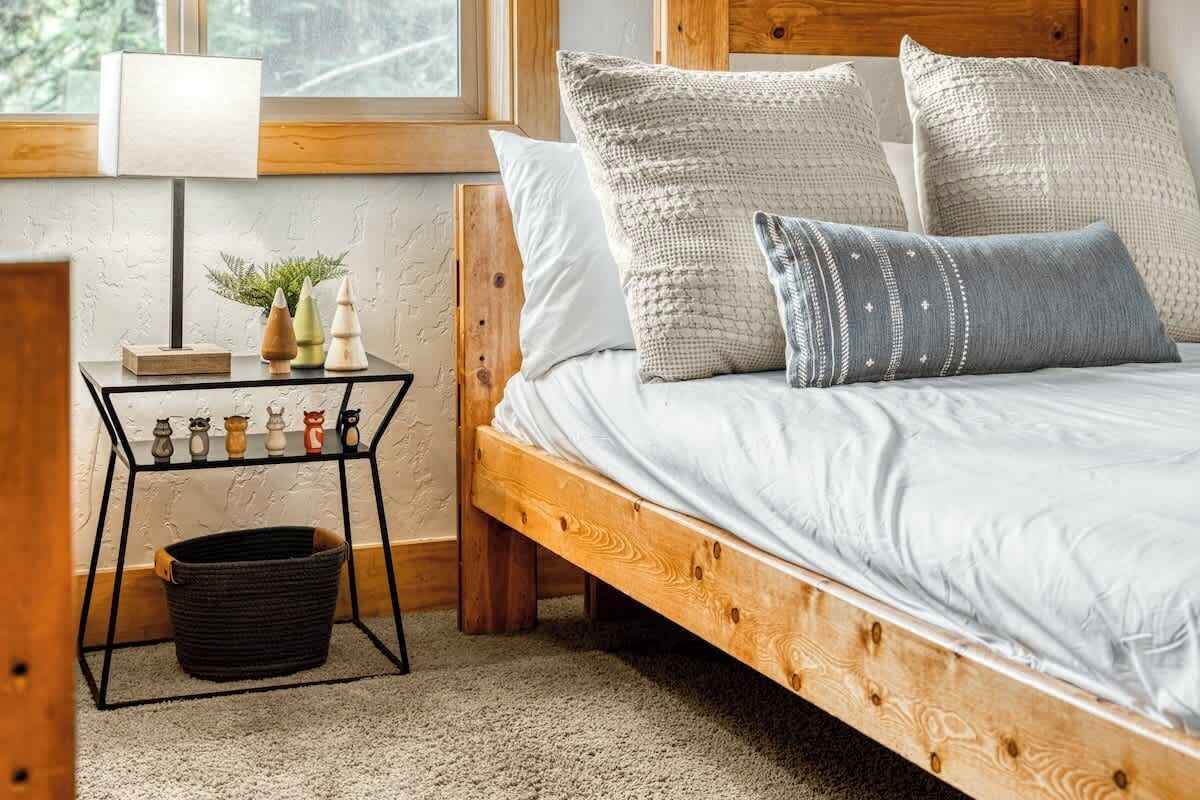 A cozy bedroom with a wooden bed, a small bedside table with decorative items, and a window with a view of trees.
