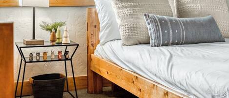 A cozy bedroom with a wooden bed, a small bedside table with decorative items, and a window with a view of trees.