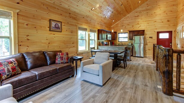Un interior de cabaña espacioso con techo de madera, comedor, cocina con electrodomésticos modernos y una zona de estar acogedora con sofá y sillones de cuero.