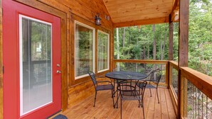 Una cabaña de madera con puerta roja, un porche cubierto con mesa y sillas, y ventanas amplias con vistas a un bosque.