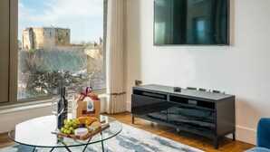 Ein modernes Wohnzimmer mit großem Fenster, einem Flachbildfernseher, einem Glastisch mit Flasche, Gläsern und einer Käseplatte.