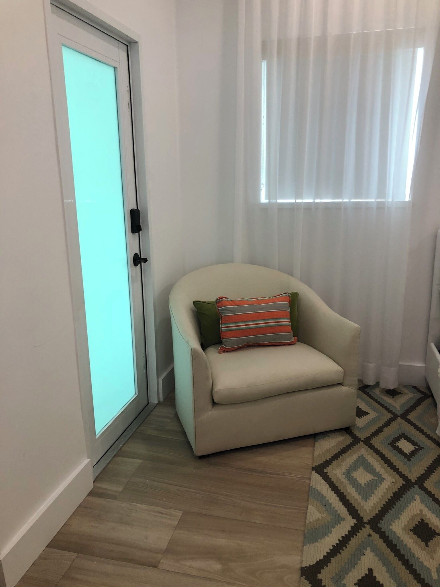 A small, cozy room with a beige armchair, a patterned rug, and a window with sheer curtains.