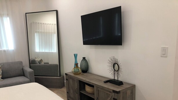 Ett modernt sovrum med en stor säng, en grå soffa, ett träbord med dekorativa föremål, en stor spegel och en väggmonterad TV.