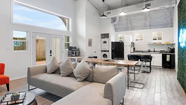 Una sala de estar moderna con un sofá gris, una mesa de comedor y una cocina con armarios blancos y un refrigerador negro.
