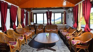 A room with a round table surrounded by wicker chairs, large windows with red curtains, and a view of mountains. - Kumaon Roop Resort (Kosiyakutoli)