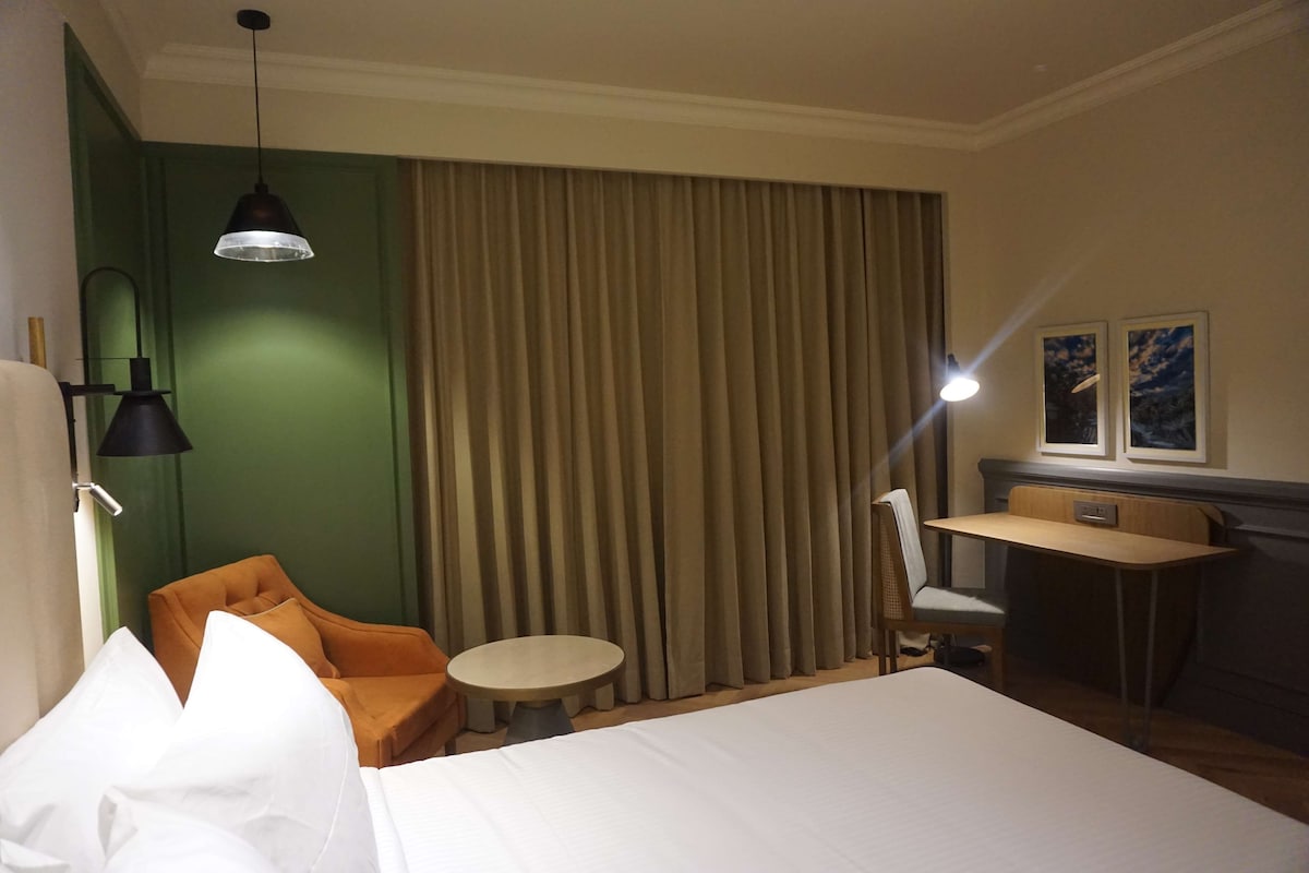 A hotel room with a bed, a round table, a chair, a desk, and a large window with curtains.