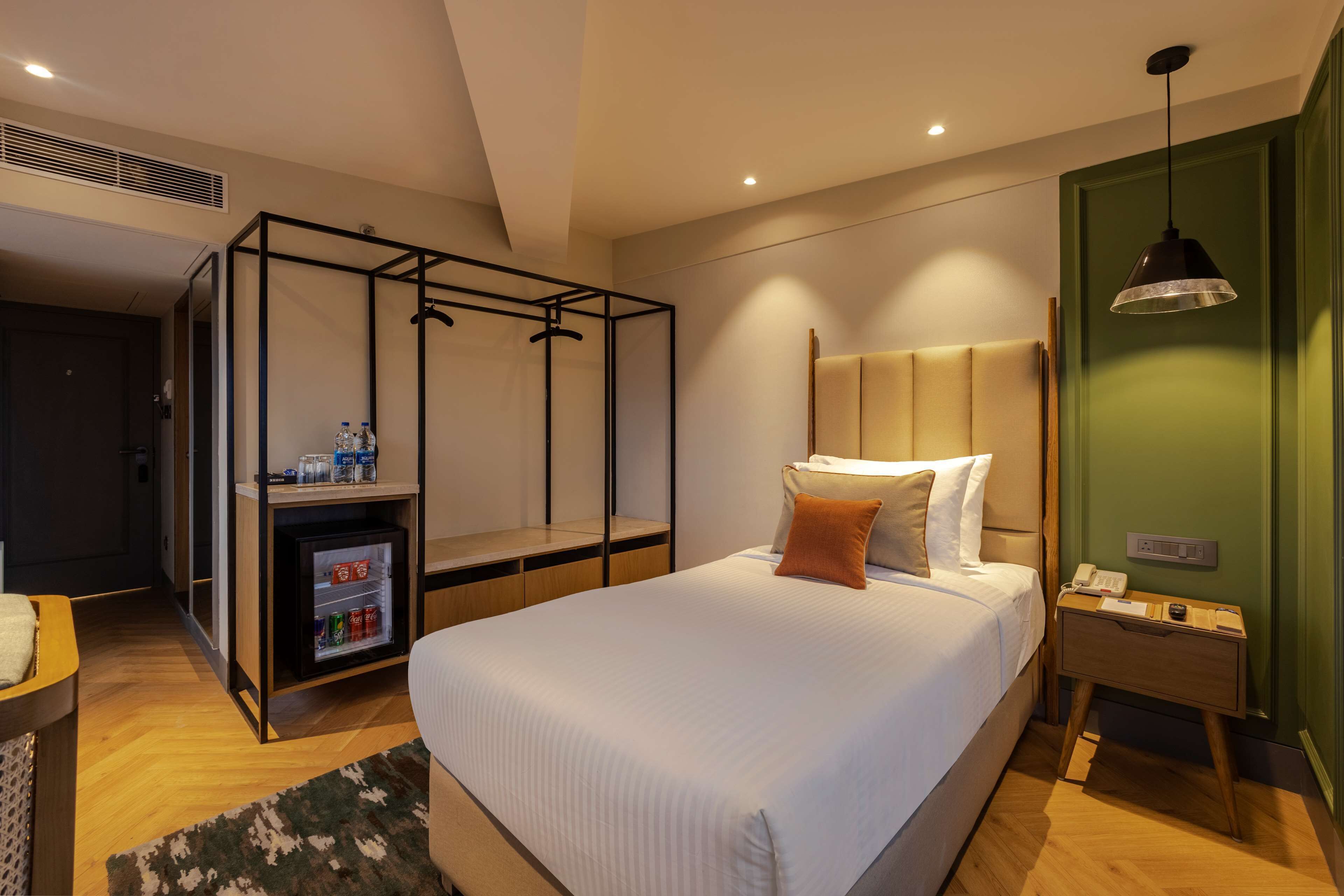 A modern hotel room with a large bed, a bedside table, a nightstand, a wardrobe, and a mini-fridge.