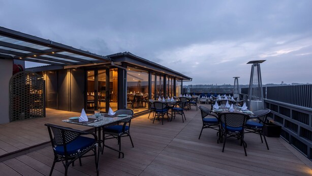 An outdoor dining area with tables and chairs, a covered section with a glass wall, and a view of the city skyline. - Sarovar Portico Srinagar (Budgam)