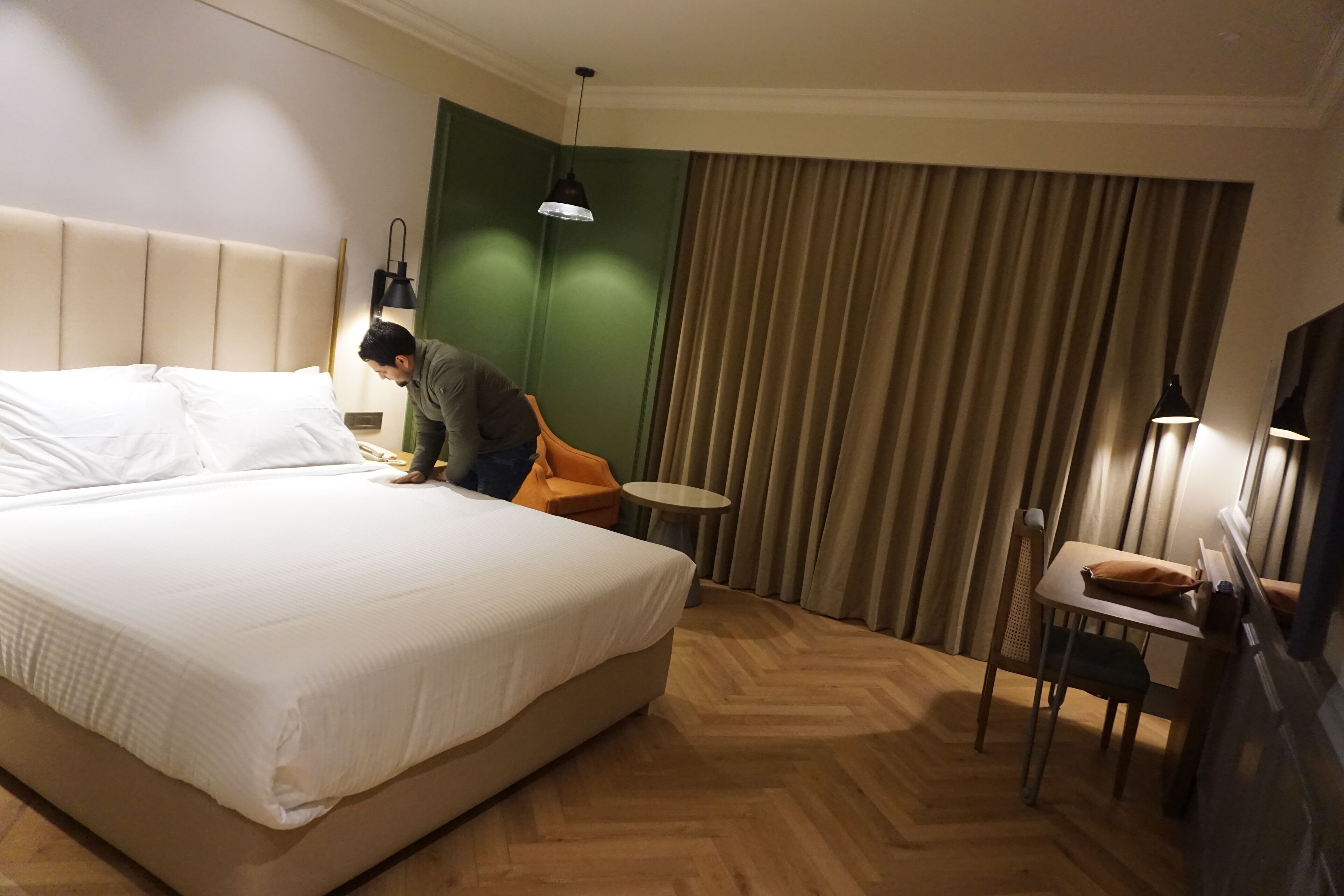 A hotel room with a large bed, a small round table, a chair, a desk with a chair, and a green accent wall.
