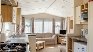 Une cuisine compacte pour camping-car, équipée d’une cuisinière, d’un évier et d’un réfrigérateur, ainsi que d’un canapé et d’un téléviseur, le tout dans un espace bien éclairé et agrémenté de rideaux.