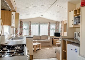 A compact RV kitchen with a stove, sink, and refrigerator, a sofa, and a television in a well-lit space with curtains.