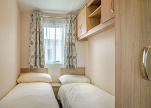 Una habitación pequeña y luminosa con dos camas, una ventana con cortinas traslúcidas y armarios de madera.