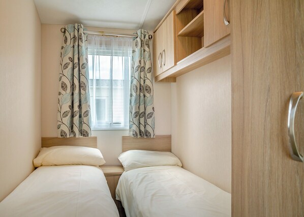 A small, well-lit room with two beds, a window with sheer curtains, and wooden cabinets.