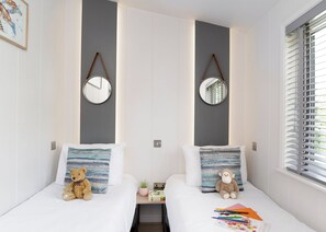 Une chambre à deux lits, chacun étant agrémenté d’un ours en peluche, d’une petite table de chevet, d’un miroir et d’une fenêtre avec des stores.
