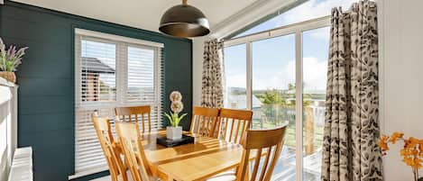 A dining area with a wooden table and chairs, a hanging light fixture, and a view through large windows.