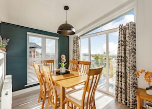 Un espace repas comprenant une table et des chaises en bois, un luminaire suspendu et une vue dégagée sur l’extérieur grâce à de grandes fenêtres.