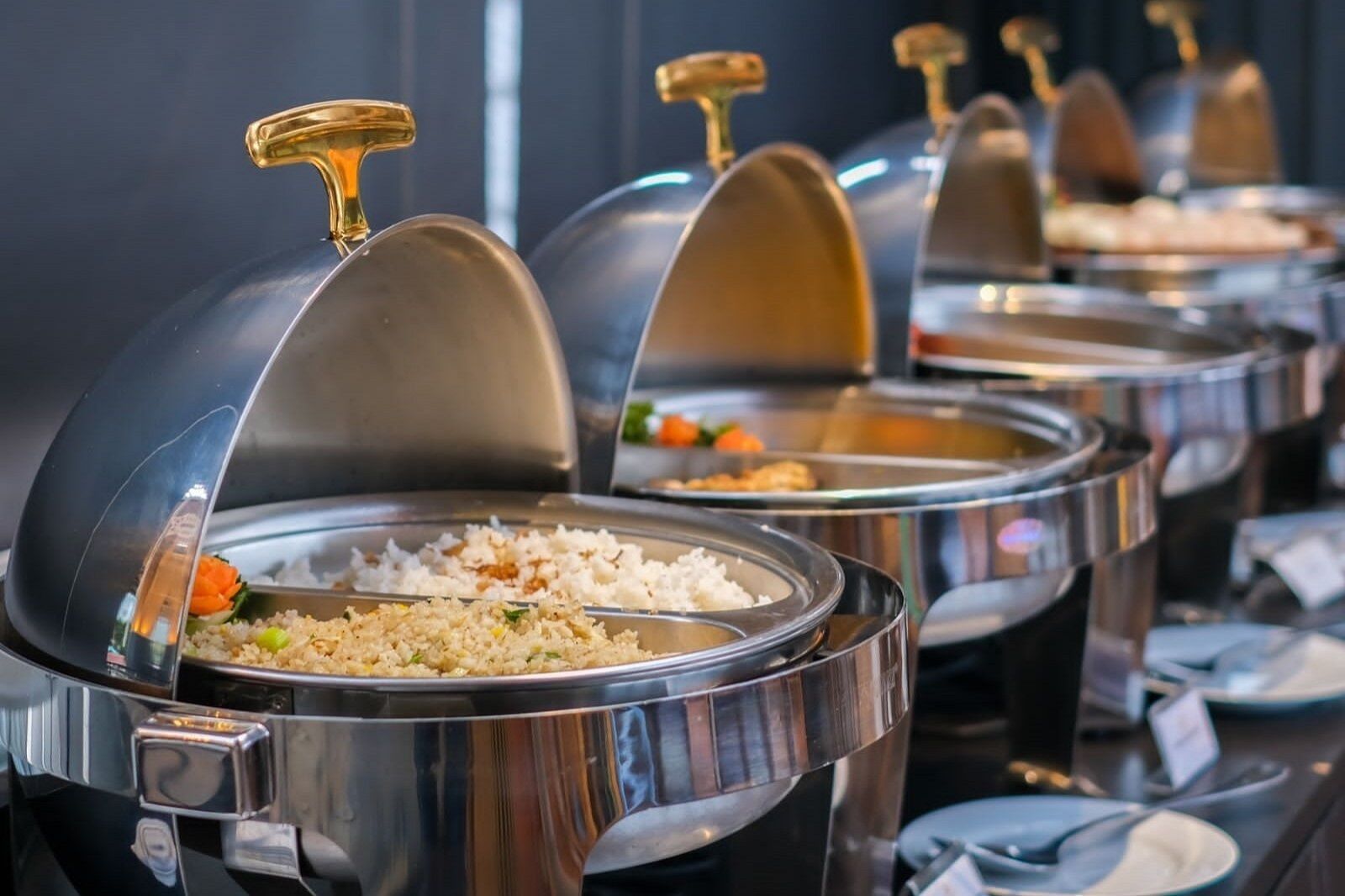 A buffet with multiple food stations, each covered with a metallic dome and labeled with a gold-colored handle.
