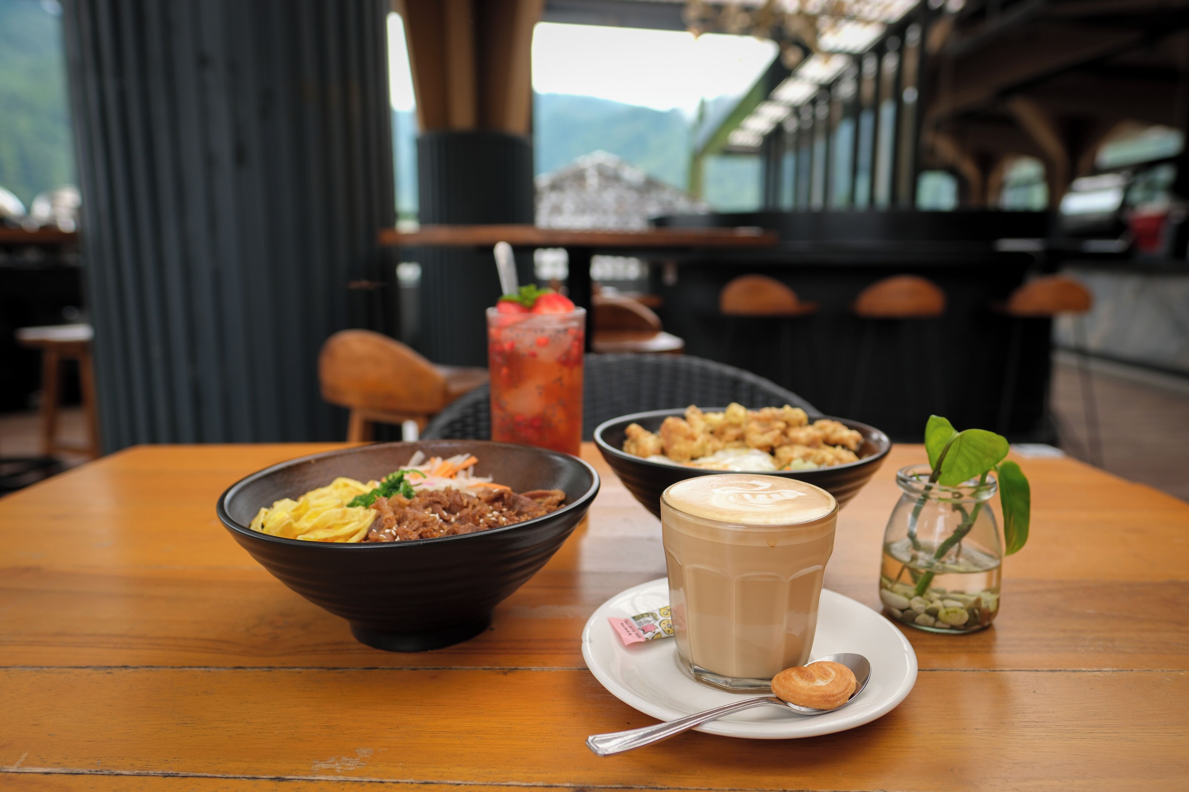 A wooden table with a bowl of food, a glass of iced coffee, and a small vase with a plant.