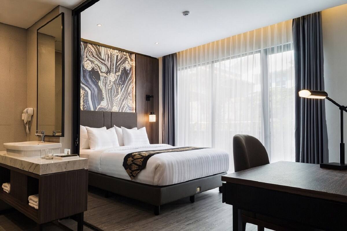 A hotel room with a large bed, a desk, a vanity with a mirror, and a large window with curtains.