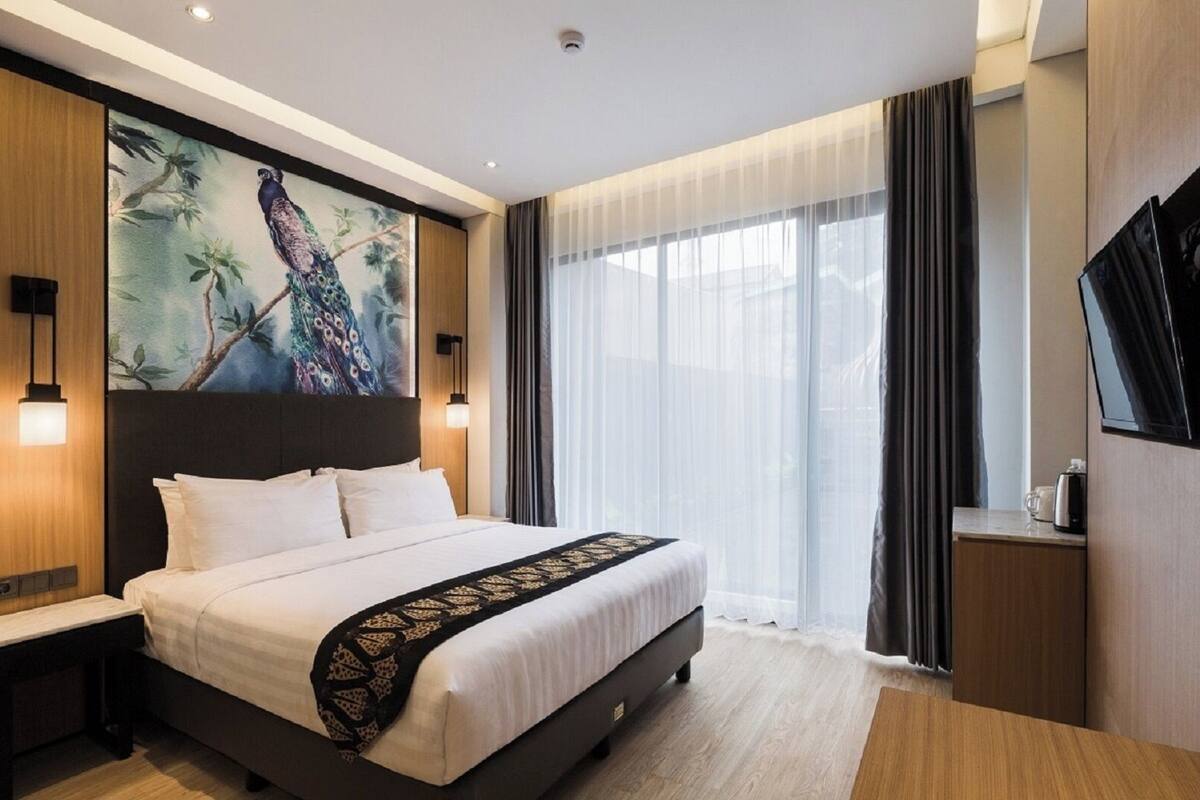 A modern hotel room with a large bed, a painting of a peacock, a TV, and a window with curtains.
