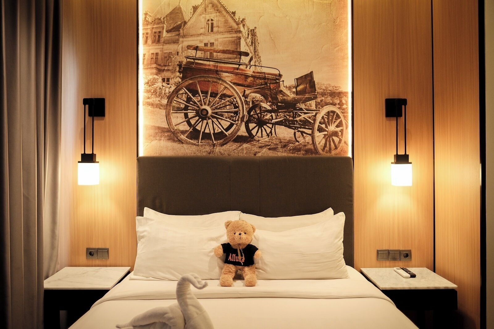 A hotel room with a bed, a teddy bear on the pillow, a remote control on the nightstand, and a wall-mounted picture of a carriage.