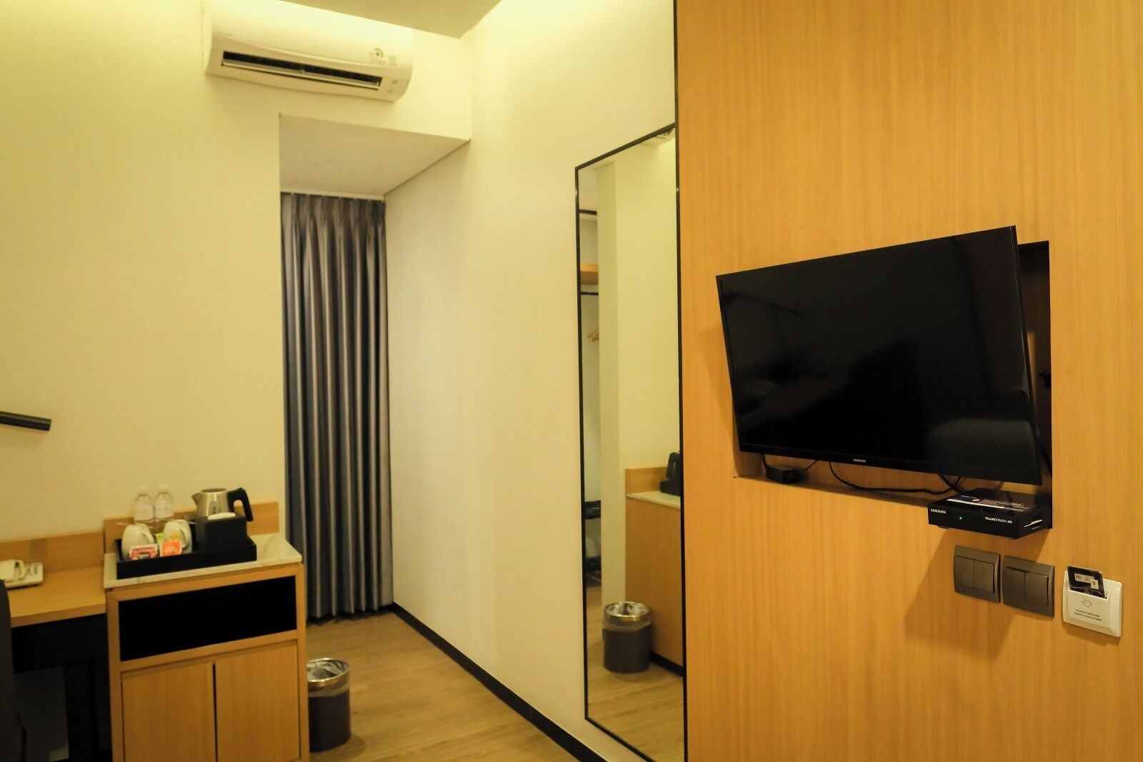A hotel room with a flat-screen TV mounted on a wooden wall, a mirror, a small wooden cabinet with toiletries, and a trash bin.