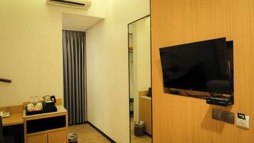 A hotel room with a flat-screen TV mounted on a wooden wall, a mirror, a small wooden cabinet with toiletries, and a trash bin.