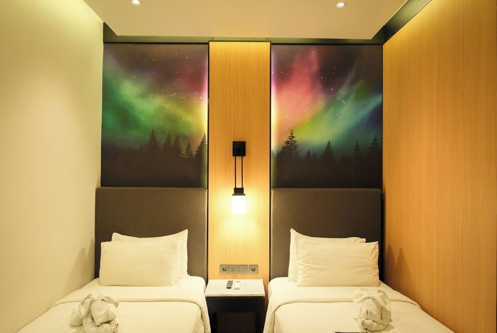 A hotel room with two beds, a nightstand, a wall-mounted lamp, and a mural of the aurora borealis.
