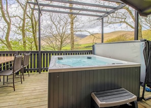 Een hottub op een houten terras, met stoelen en een tafel, omringd door een houten omheining en bomen.