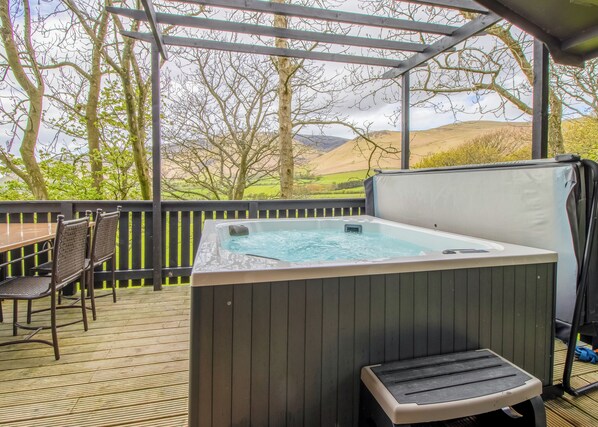 A hot tub on a wooden deck with chairs and a table, surrounded by a wooden fence and trees.