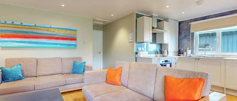 Uma sala de estar moderna com sofá e mesa de centro, uma cozinha com armários e janela, e um quadro abstrato e colorido na parede.
