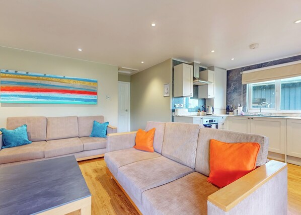 A modern living room with a sofa and a coffee table, a kitchen with cabinets and a window, and a colorful abstract painting on the wall.
