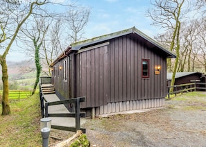 Een houten gebouw in landelijke stijl met een trap naar een deur, omgeven door bomen en een omheining.