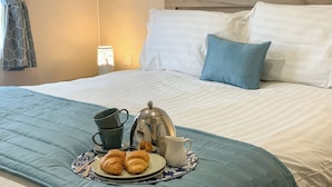 Un letto rifatto con un copriletto blu, un piatto con croissant e una caffettiera, e una lampada sullo sfondo.