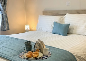 Una cama bien hecha con una manta azul, un plato con croissants y una cafetera, y una lámpara al fondo.