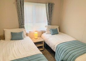 A bedroom with two single beds, a bedside table with a lamp, a window with curtains, and a view of the outside.