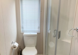 Un baño con inodoro, un rollo de papel higiénico, cabina de ducha de vidrio y una ventana con persianas.