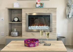 A modern living room with a fireplace, a clock, a shelf with books, and a table with a candy bowl and glasses.