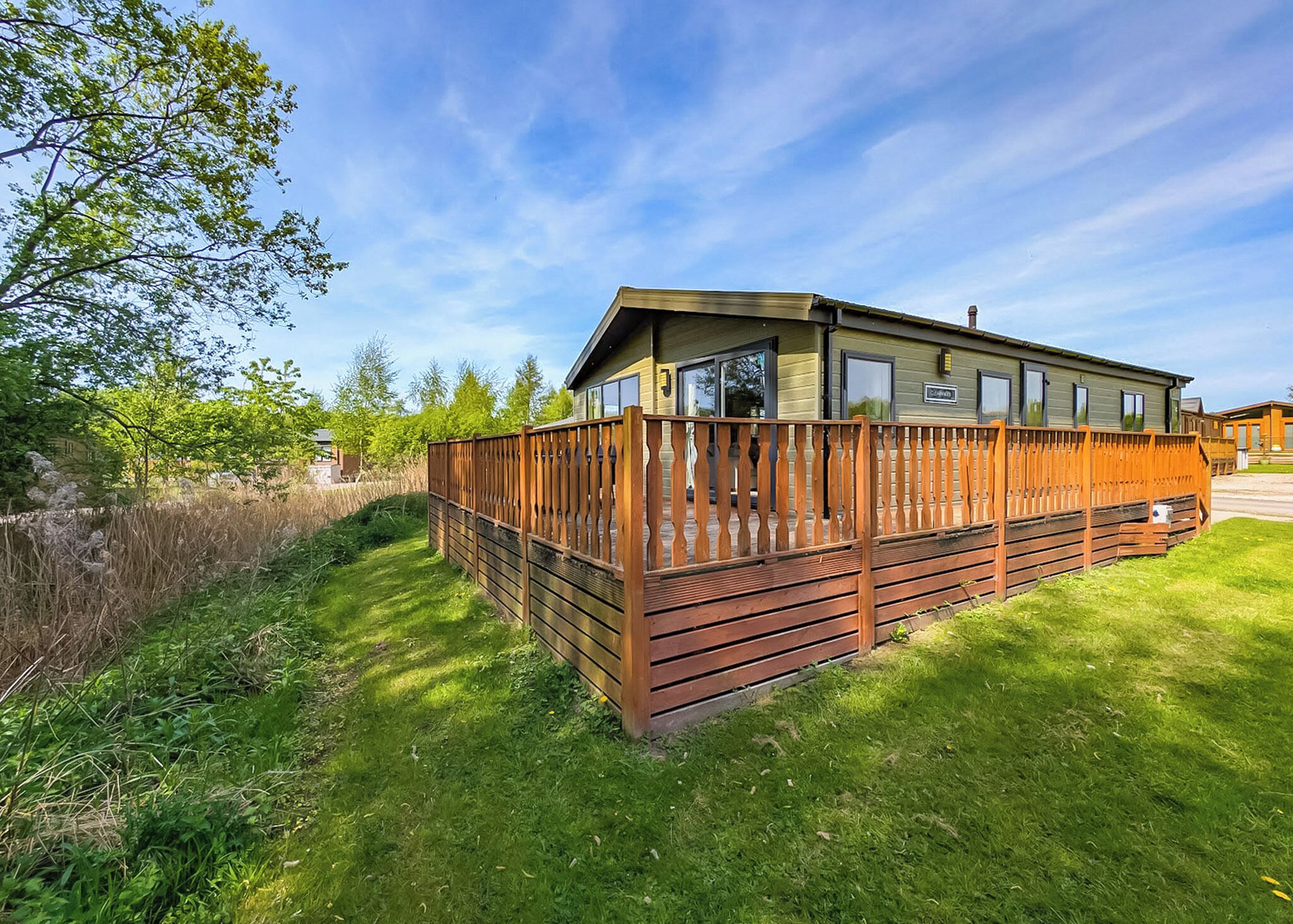 A wooden decked RV with a covered porch, surrounded by lush greenery and a clear sky.