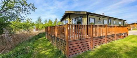 A wooden decked RV with a covered porch, surrounded by lush greenery and a clear sky.