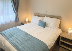 Un letto rifatto con cura, con una coperta azzurra, cuscini bianchi e una testiera in legno. Sono presenti due comodini con lampade, e una finestra con tende leggere.